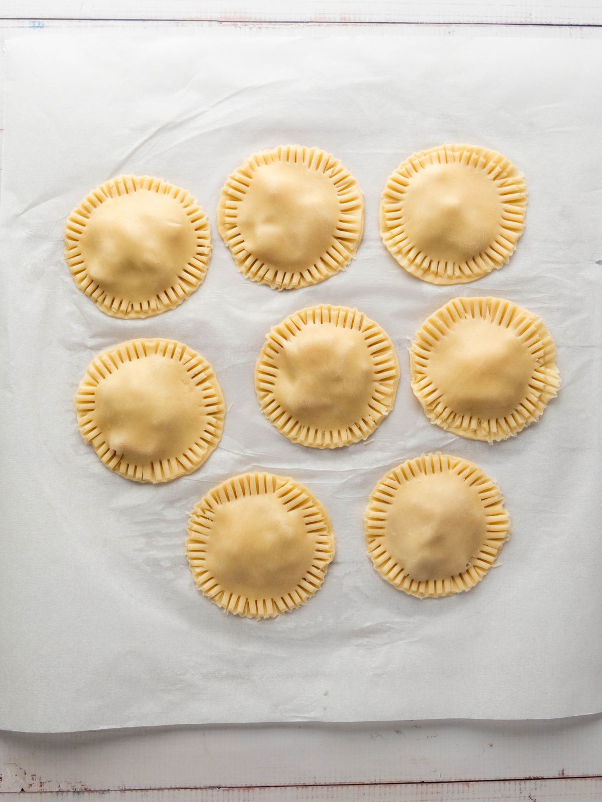 Seven raw, circular hand pies with crimped edges arranged on parchment paper.