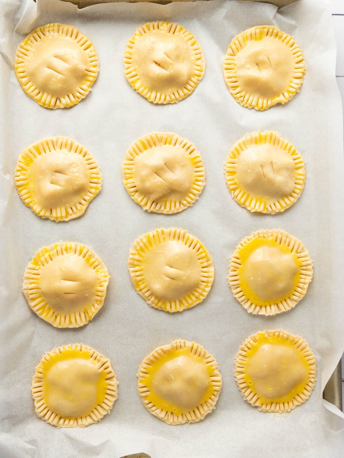 Twelve round unbaked hand pies arranged in a grid on a parchment-lined baking sheet.