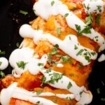 Two beef and bean burritos are topped with red sauce, a drizzle of white cream, and fresh chopped cilantro on a black plate. A lime wedge is visible at the top edge of the image.
