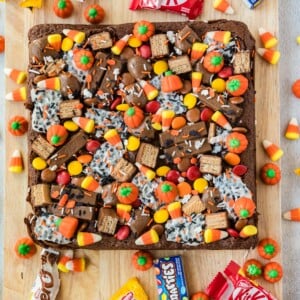 A square brownie topped with candy corn, pumpkin-shaped candies, chocolate bars, and colorful sprinkles, sitting on a wooden board.