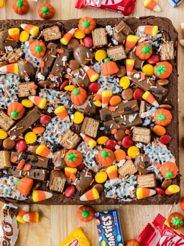 A square brownie topped with candy corn, pumpkin-shaped candies, chocolate bars, and colorful sprinkles, sitting on a wooden board.