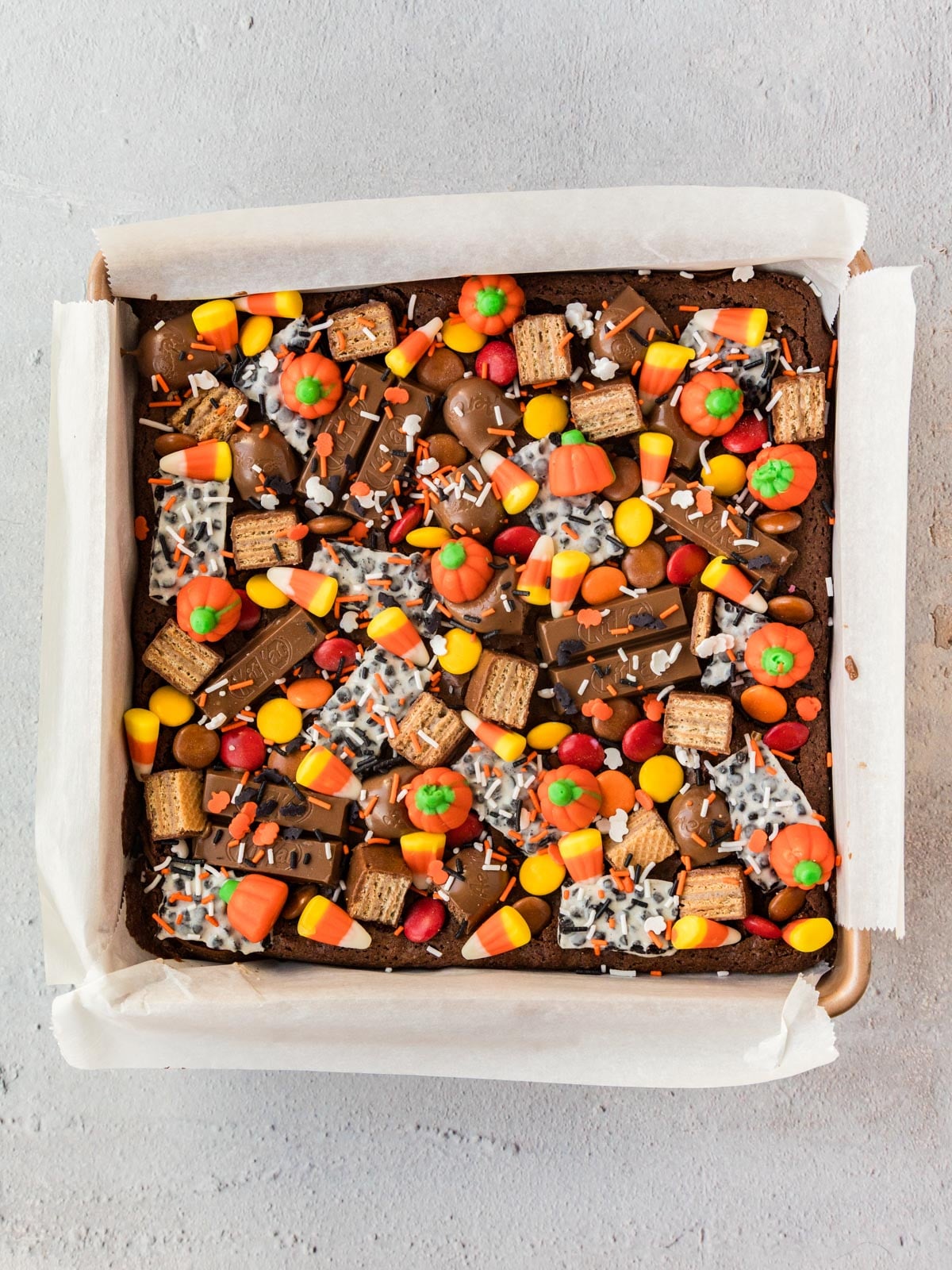 A square brownie topped with candy corn, chocolate-covered wafer bars, pumpkin-shaped candies, and sprinkles in a parchment-lined baking pan.