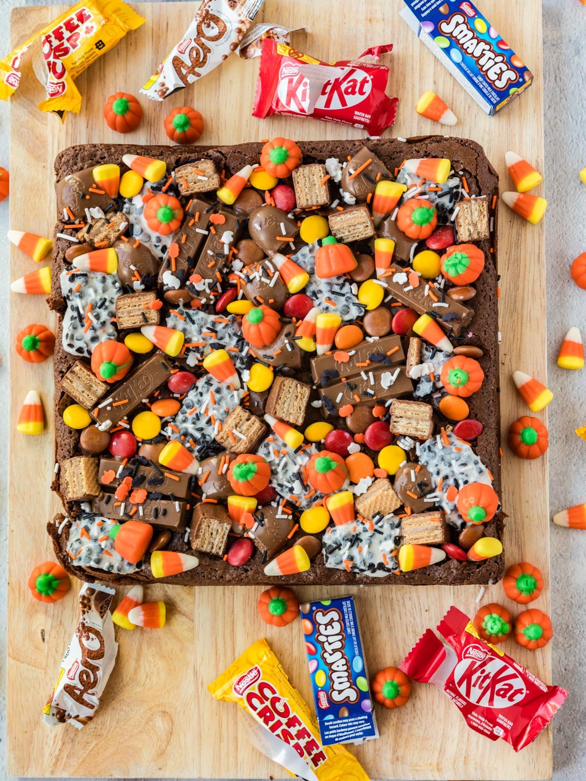 A large square brownie topped with candy pumpkins, wafer cookies, and sprinkles, surrounded by scattered Coffee Crisp, Aero, Smarties, KitKat, and candy corn on a wooden board.