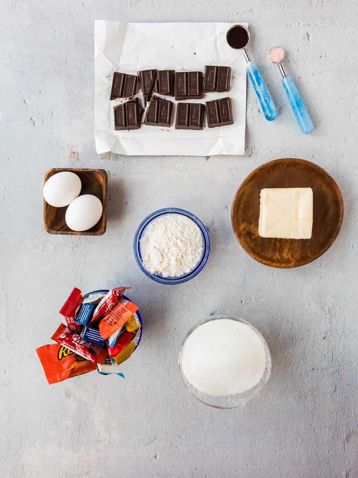 Ingredients for baking arranged on a grey surface, including chocolate squares, eggs, butter, flour, sugar, and a bowl of assorted candy bars.