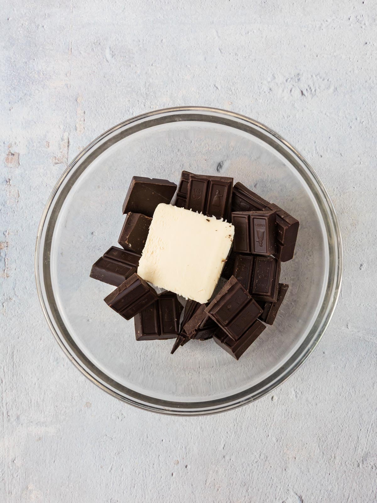 A glass bowl containing a stick of butter resting on top of several squares of dark chocolate.