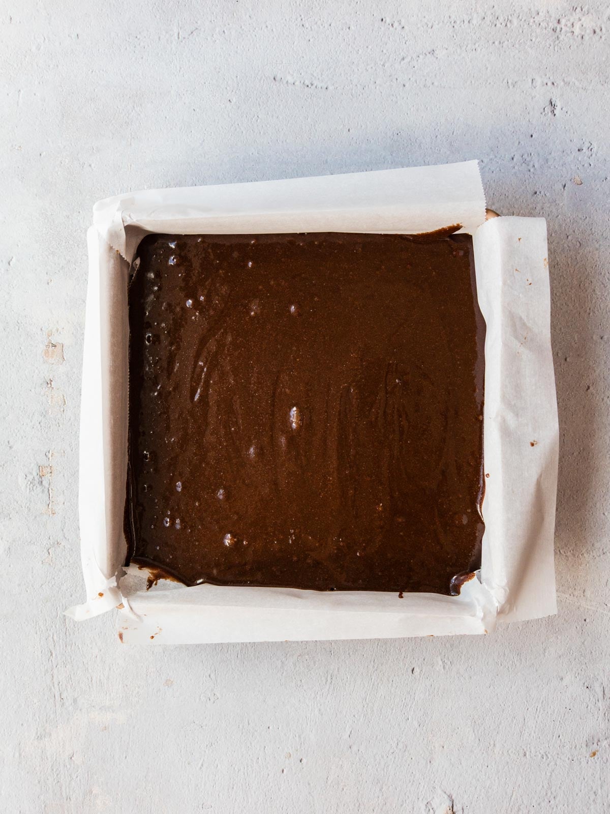 Square baking pan lined with parchment paper filled with raw brownie batter.