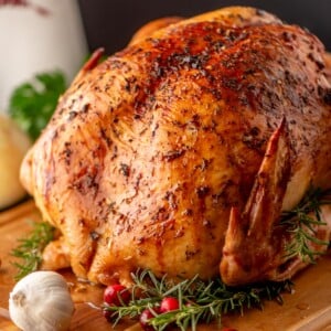A roasted chicken garnished with sprigs of rosemary, fresh cranberries, and a head of garlic on a wooden board.