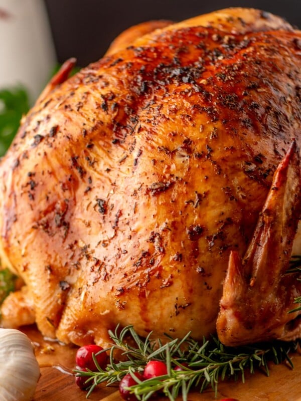 A roasted chicken garnished with sprigs of rosemary, fresh cranberries, and a head of garlic on a wooden board.