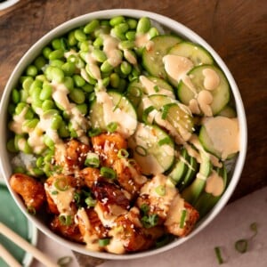 A rice bowl topped with teriyaki chicken, edamame, sliced cucumbers, and avocado, drizzled with a creamy sauce and sprinkled with sesame seeds.