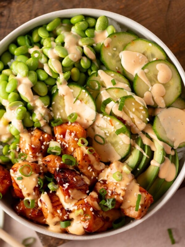 A rice bowl topped with teriyaki chicken, edamame, sliced cucumbers, and avocado, drizzled with a creamy sauce and sprinkled with sesame seeds.