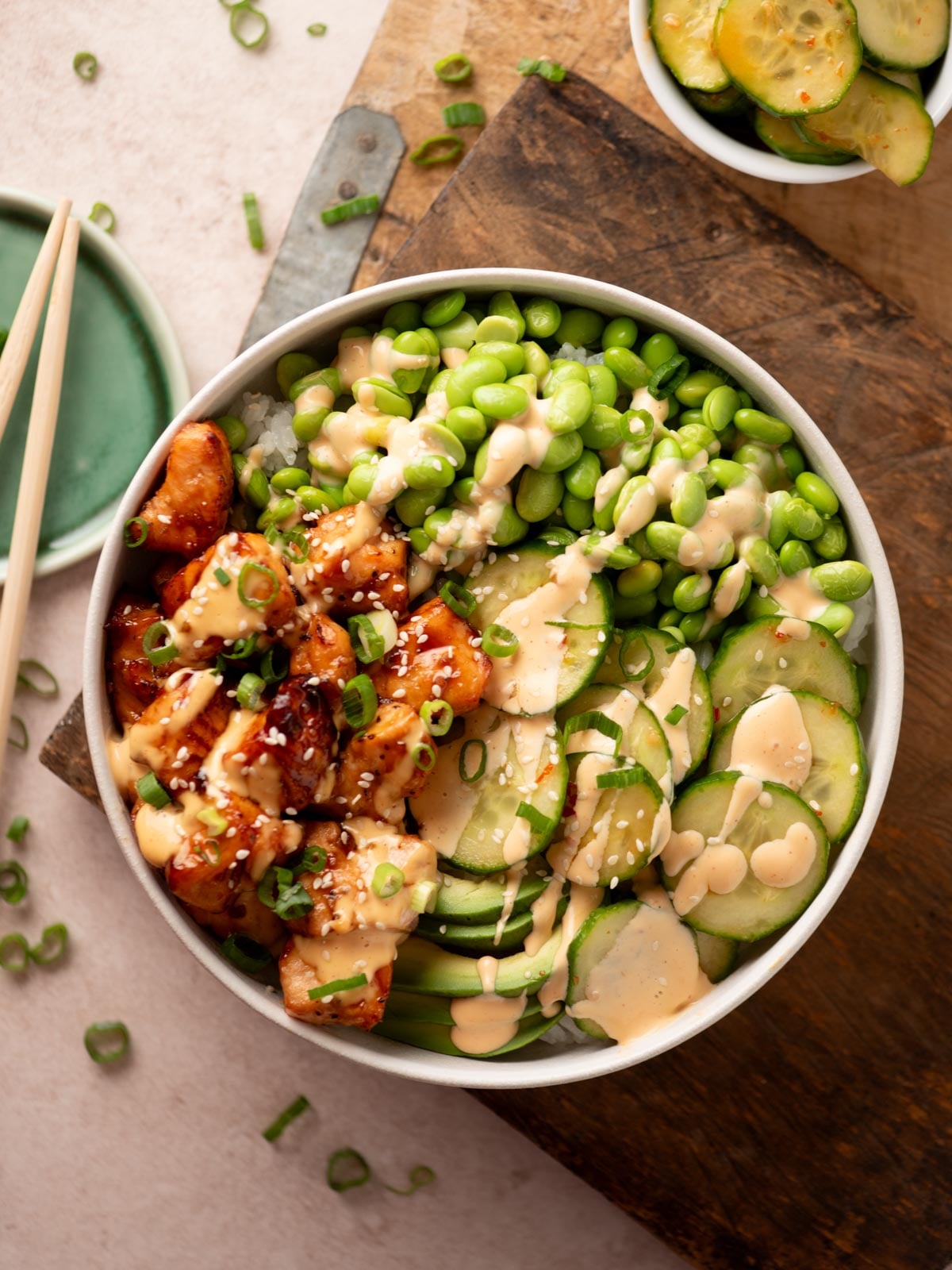 A bowl of rice topped with glazed chicken, edamame, sliced cucumbers, and avocado, drizzled with creamy sauce and garnished with sesame seeds and green onions.