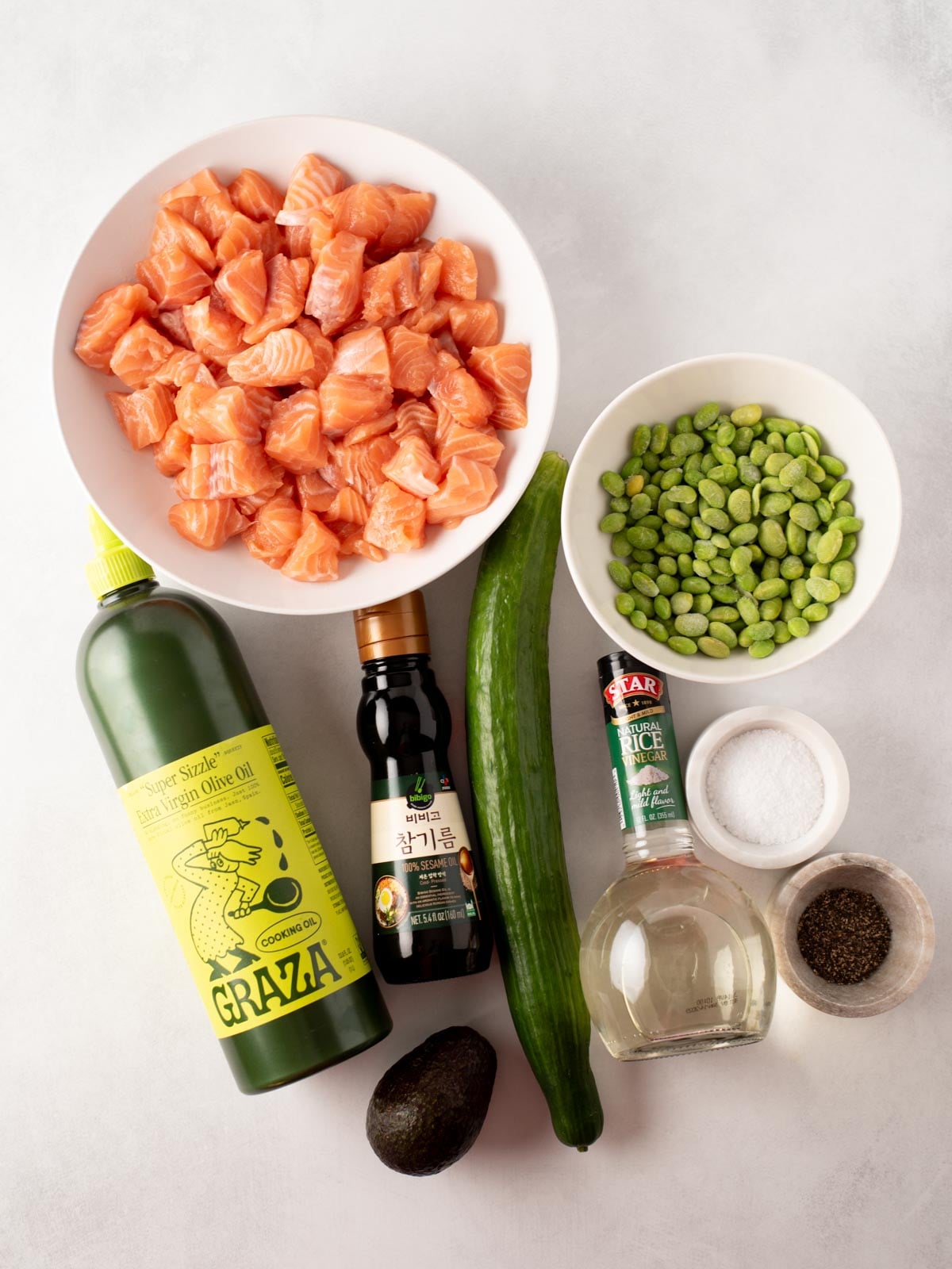 Ingredients for a poke bowl including cubed salmon, edamame, a cucumber, avocado, bottles of olive oil, sesame oil, and rice vinegar, plus small bowls of salt and pepper.