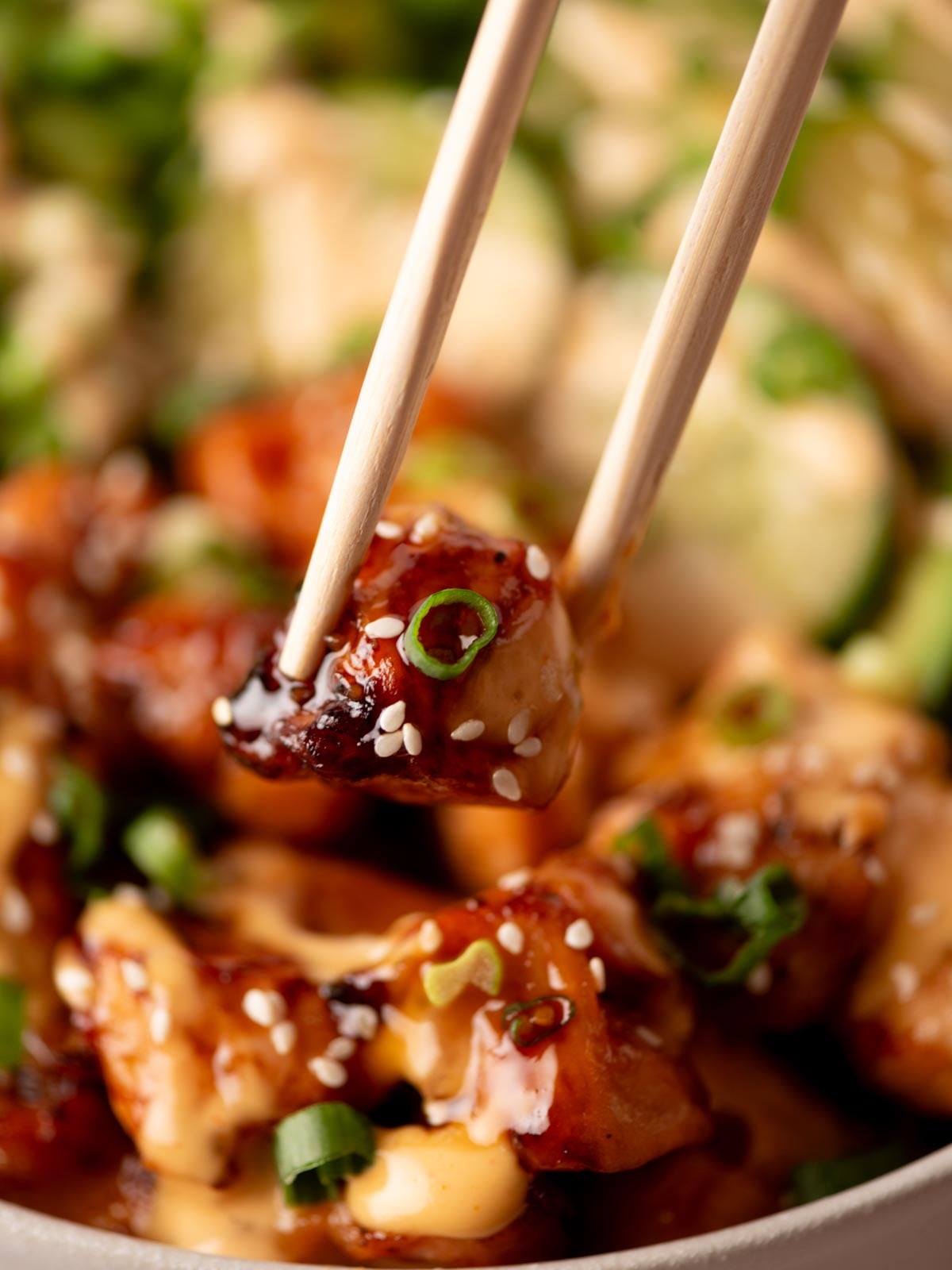 Wooden chopsticks holding a piece of glazed chicken topped with sesame seeds and green onions over a bowl of food.