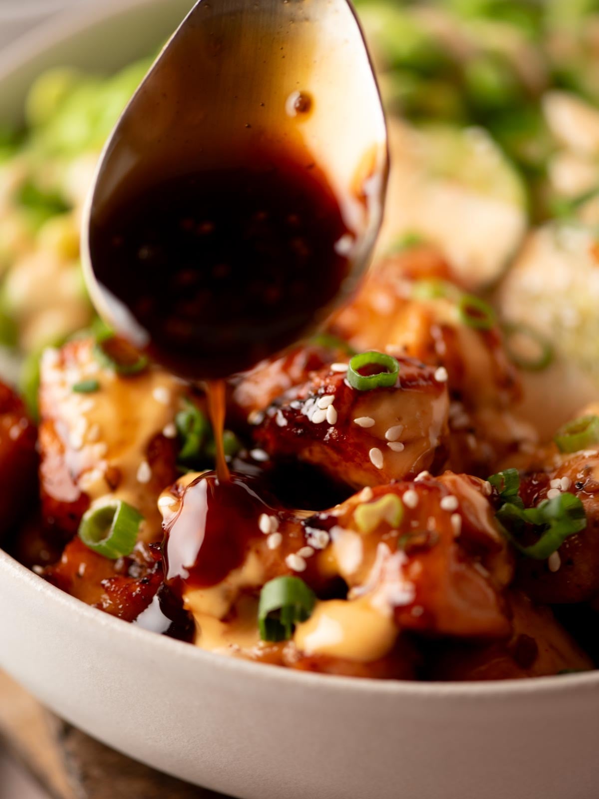 A spoonful of dark sauce drizzled over chicken bites topped with sesame seeds and green onions in a bowl.